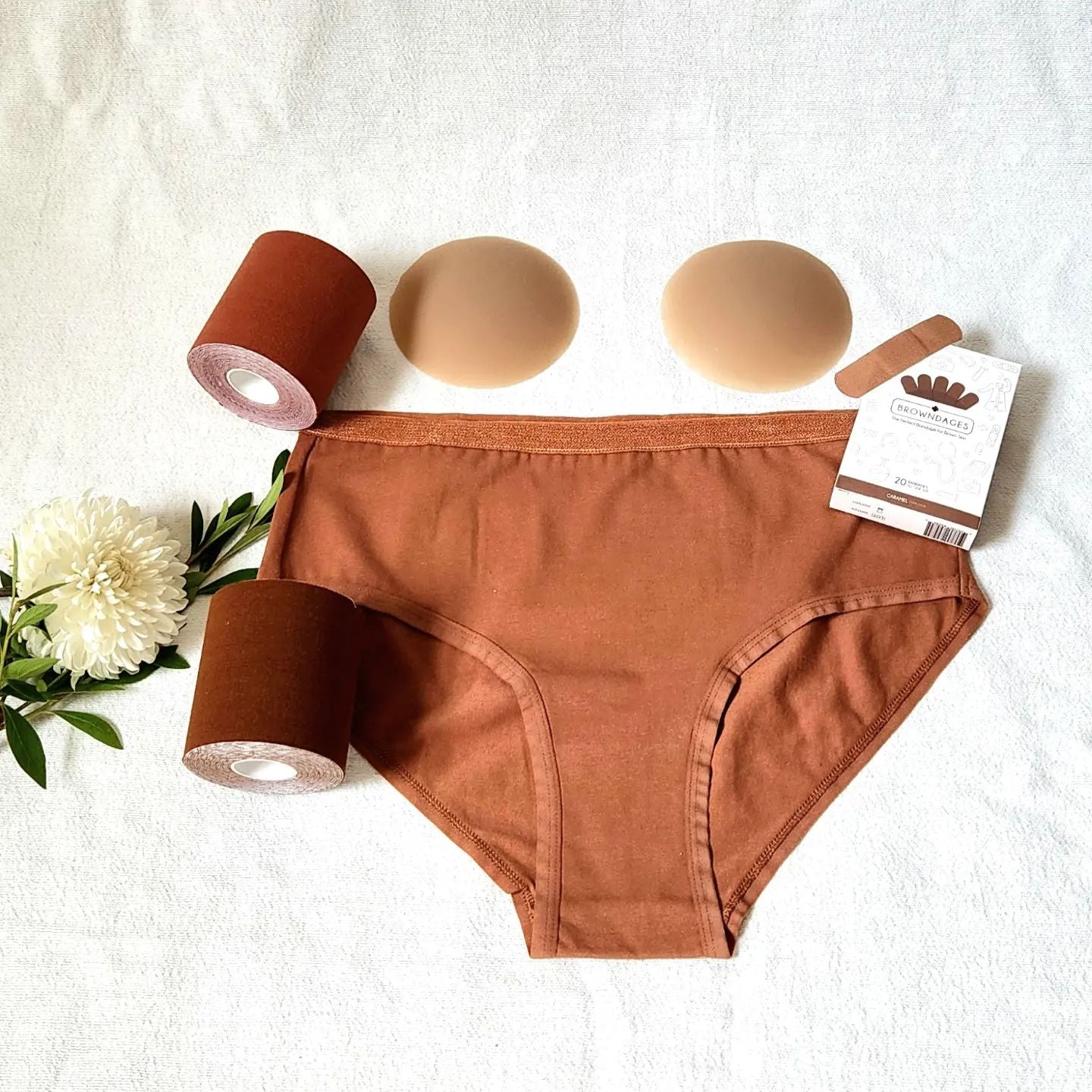 A bundle deal including a pair of medium brown women's underwear with skin tone matching nipple covers, two rolls of boob tape, and band-aids laying on a white background with a flower. Shop for your skin tone in 15 minutes or less.