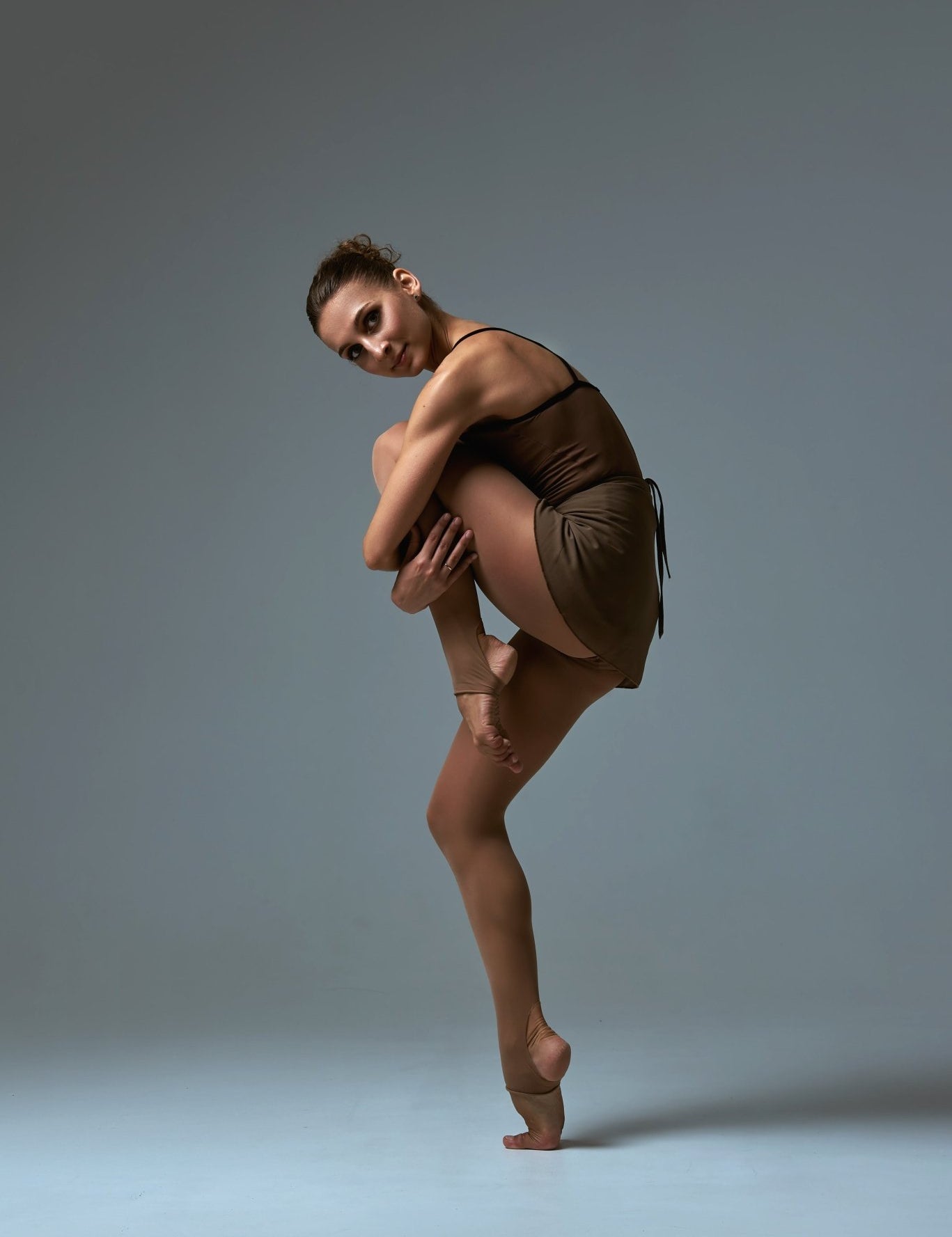 A tanned woman posing on one leg in brown stirrup dance tights.