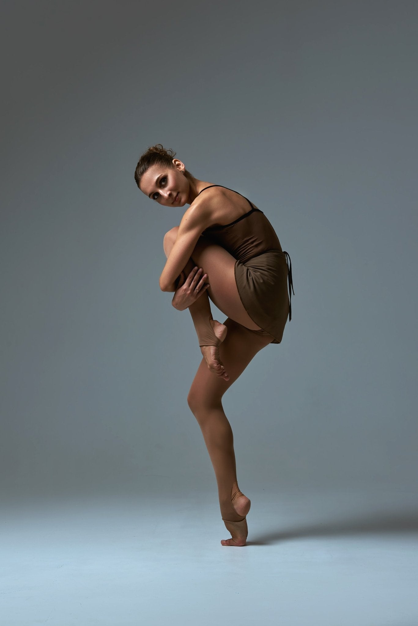 A tanned woman posing on one leg in brown stirrup dance tights.