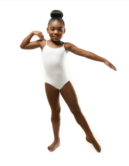 A happy young Black girl in a bun, wearing a white leotard and medium brown transition tights the color “brazen brown”. One leg is up to the knee and the other over her foot.